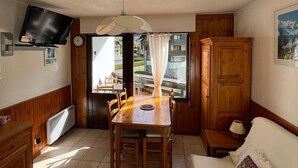 A compact room with a wooden dining table, chairs, a television mounted on the wall, a wooden cabinet, and a window with curtains. - 2-room apartment with pets allowed, balcony and garage (Samoëns)