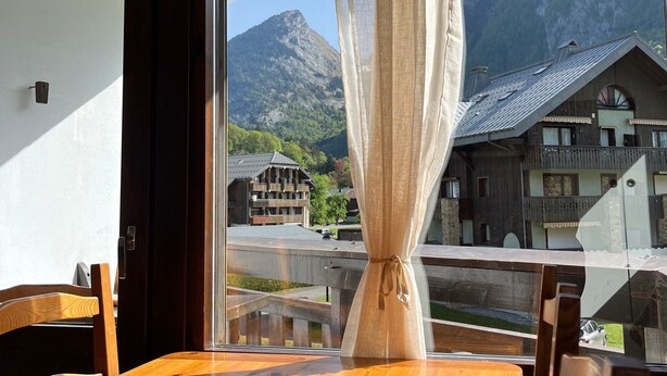 A room with a wooden table and chairs, a window with a curtain, and a view of mountain buildings and a bridge. - 2-room apartment with pets allowed, balcony and garage (Samoëns)