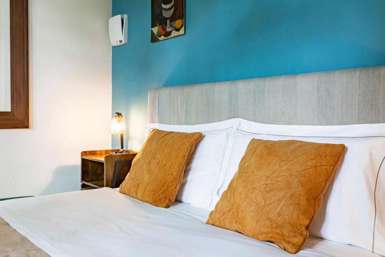 Un lit avec du linge de lit blanc et deux oreillers marron, une table de chevet avec une lampe, et un mur bleu orné d’un tableau.