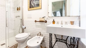 A bathroom with a glass shower enclosure, a bidet, a toilet, a sink with a vintage-style base, and a framed artwork on the wall.