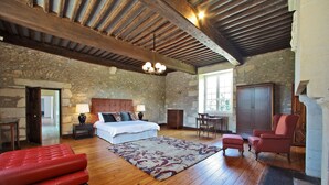 A bedroom with a stone wall, wooden ceiling beams, a bed with a headboard, a red sofa, a patterned rug, and a wooden cabinet. - Stunning chateau, built in 1611, beautifully renovated, furnished and equipped. (Saint-Geyrac)