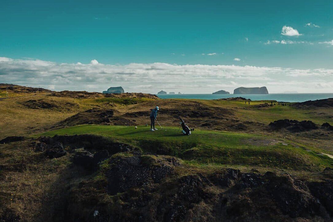 A golf course with a person standing and another preparing to take a shot, surrounded by rocky terrain and distant islands.