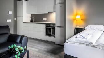 A modern hotel room with a kitchenette, a black leather sofa, a small coffee table with a plant, and a bedside lamp.