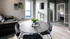 A modern living room with a grey sofa, a round dining table with chairs, a TV mounted on the wall, and a small plant on a shelf. - The New Post Office (Vestmannaeyjar)