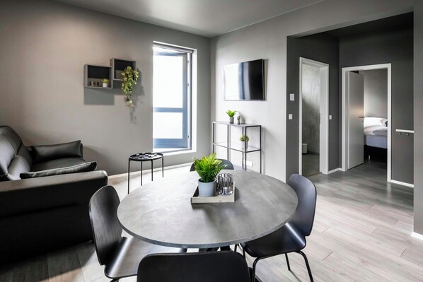 A modern living room with a grey sofa, a round dining table with chairs, a TV mounted on the wall, and a small plant on a shelf. - The New Post Office (Vestmannaeyjar)