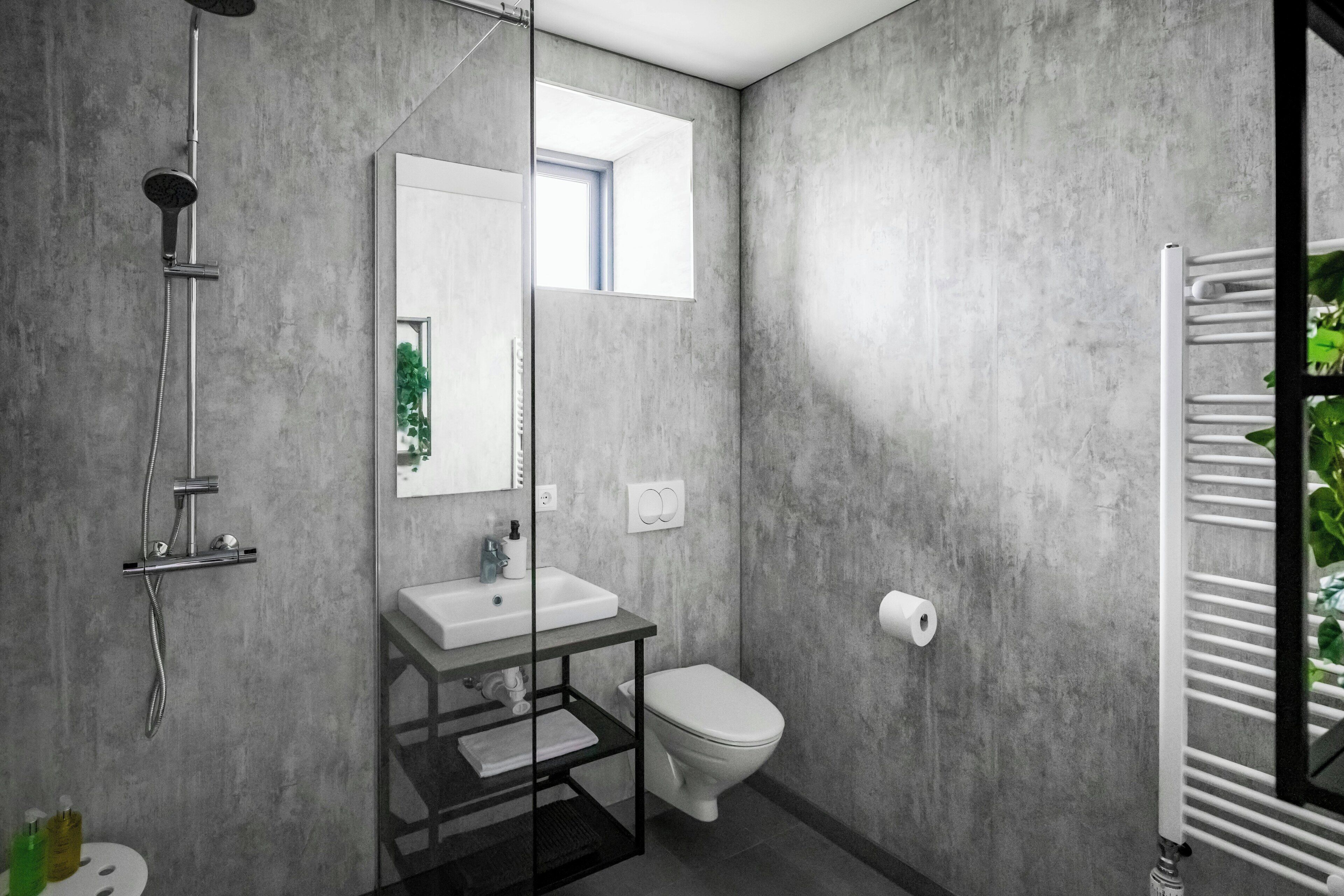 A modern bathroom with a concrete wall, a glass shower enclosure, a white sink, and a toilet.