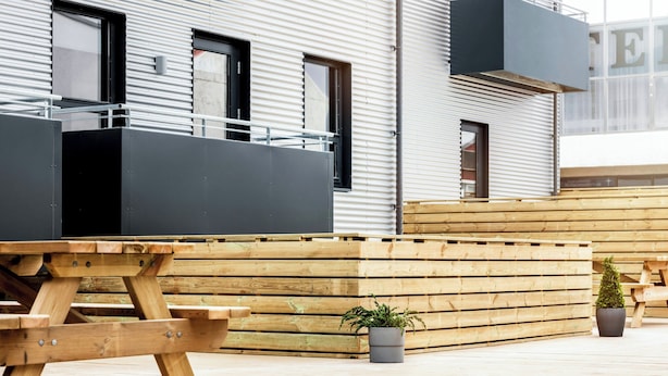 A wooden deck with a picnic table and potted plants, set against a modern building with large windows and balconies. - The New Post Office (Vestmannaeyjar)