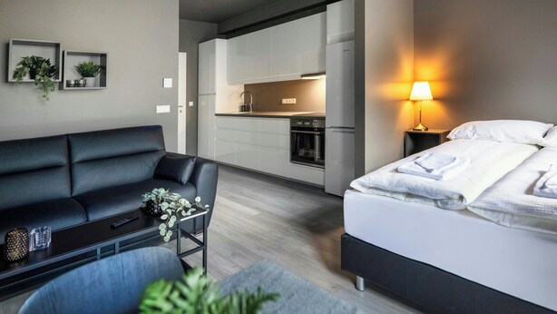 A modern hotel room with a black leather sofa, a bed with white linens, a small table with a plant, and a kitchen area in the background. - The New Post Office (Vestmannaeyjar)