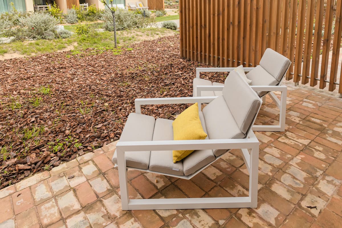 A patio with two white lounge chairs, a wooden fence, and a building with balconies in the background.