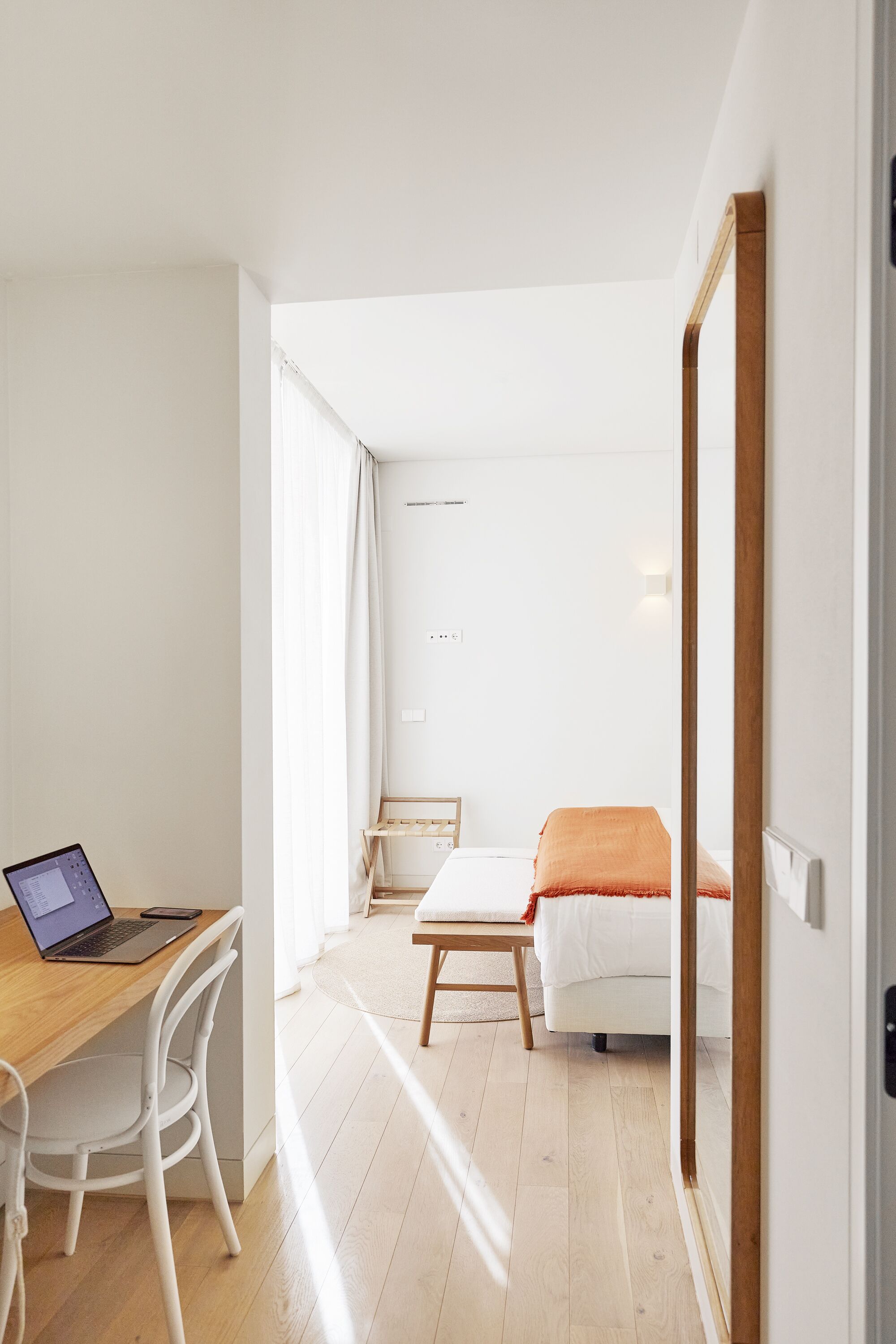 Une pièce moderne et bien éclairée, avec un bureau en bois, un ordinateur portable, une chaise blanche, un lit recouvert d’une couverture orange et un miroir à cadre en bois.