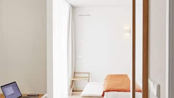 Une pièce moderne et bien éclairée, avec un bureau en bois, un ordinateur portable, une chaise blanche, un lit recouvert d’une couverture orange et un miroir à cadre en bois.