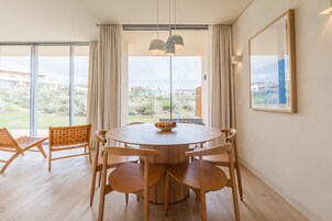 Un comedor con una mesa redonda, seis sillas de madera, un frutero y vistas a un jardín a través de ventanas amplias.