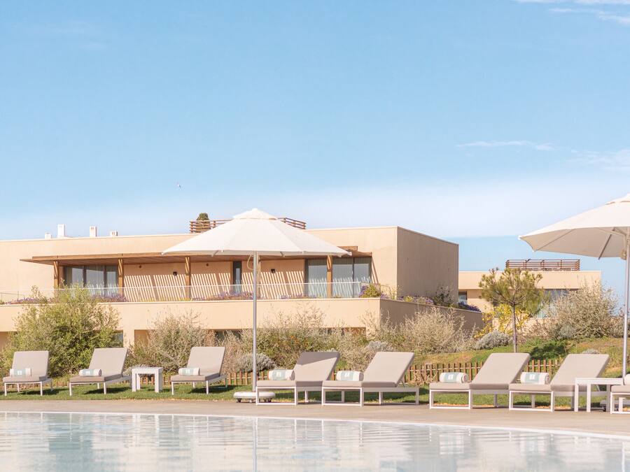 Un espace aménagé au bord de la piscine, avec des chaises longues et des parasols, un bâtiment moderne doté de balcons, et un ciel dégagé.