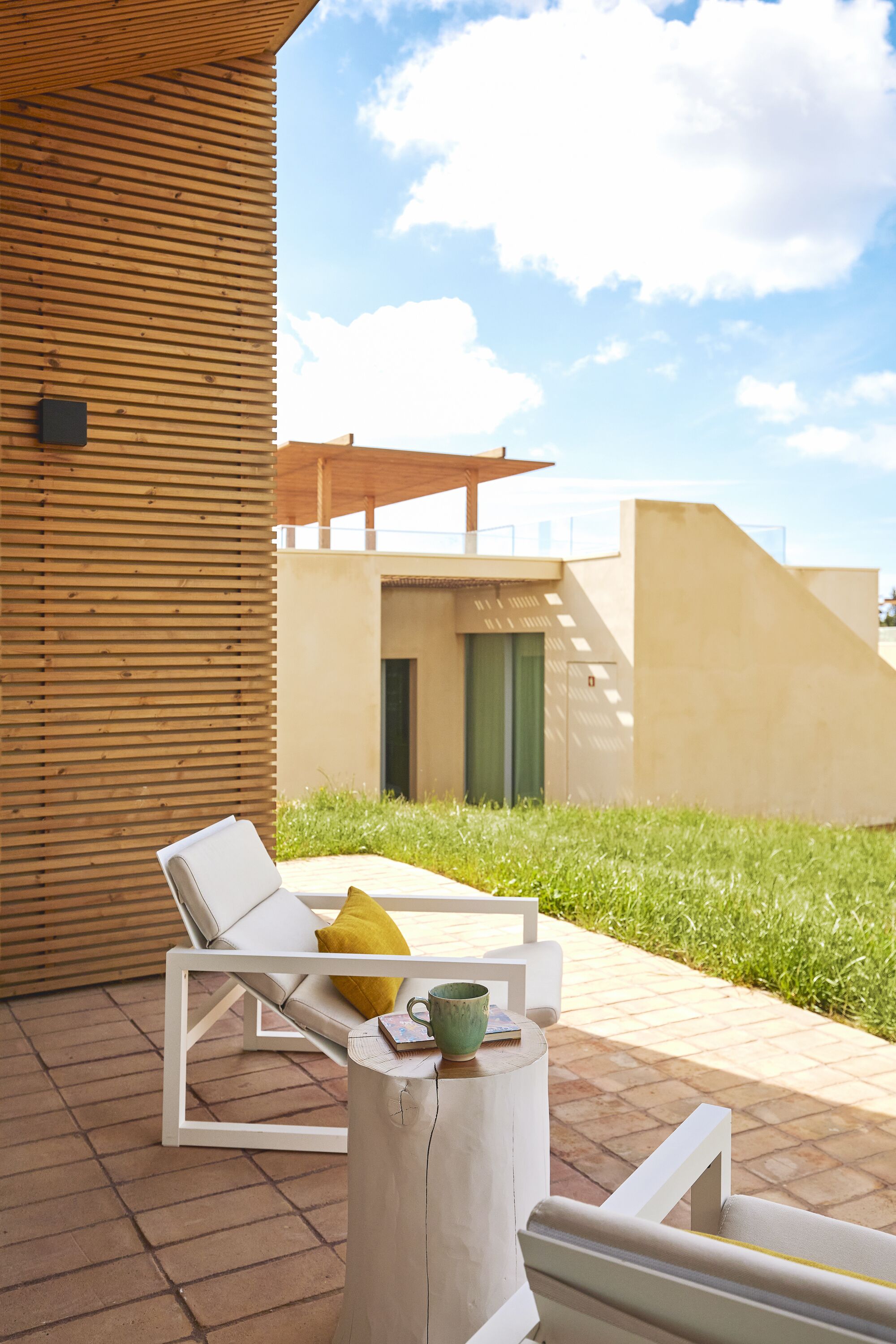 Een modern buitenverblijf met witte loungestoelen, een ronde houten tafel, een groene deur en een groene grasachtige achtergrond.