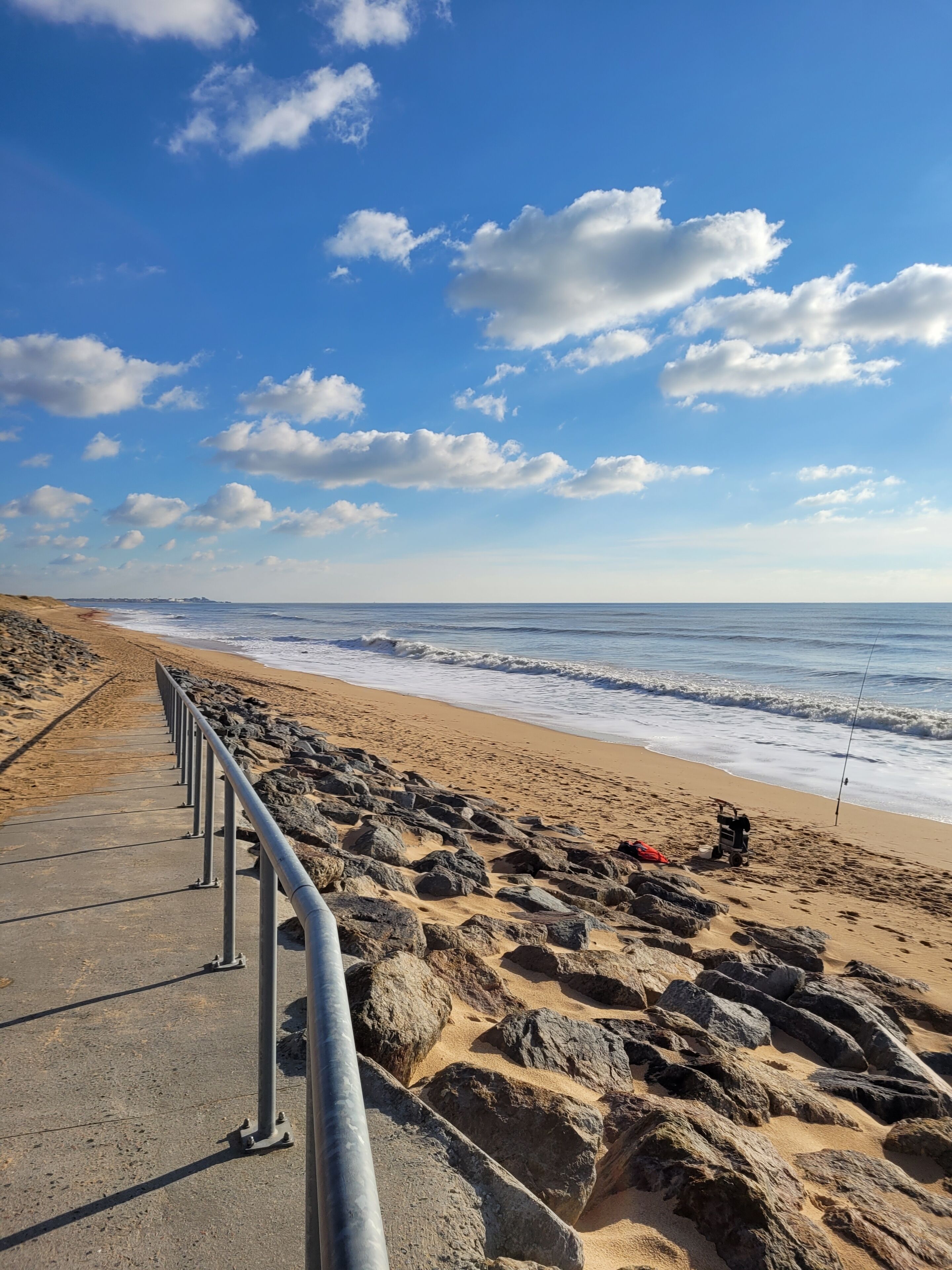 Une promenade en bord de mer, dotée d’une rambarde en métal, qui mène à une plage de sable avec de gros rochers et une canne à pêche.