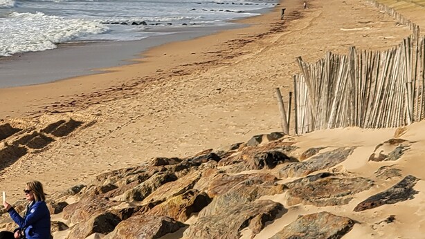 Una playa con primer plano rocoso, una valla de madera y una persona sentada sobre las rocas.