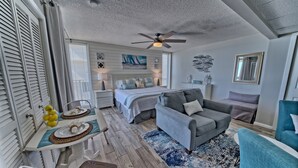 A bedroom with a bed, a gray sofa, a dining table with a chair, and a ceiling fan.