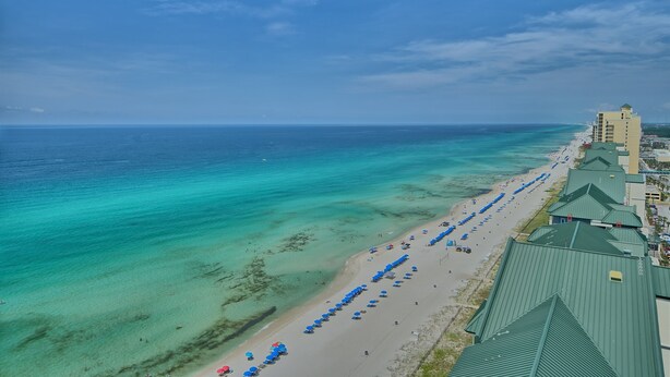 A coastal hotel with a beach view, rows of blue umbrellas, and a clear blue sky.