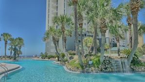 A swimming pool with a waterfall feature, surrounded by palm trees and a high-rise building.