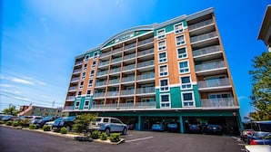 Ein mehrgeschossiges Hotel mit grün-beiger Fassade, mehreren Balkonen und einem Parkplatz davor.