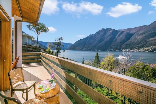 A balcony with wicker furniture, a table with a drink and flowers, and a view of a lake and mountains.