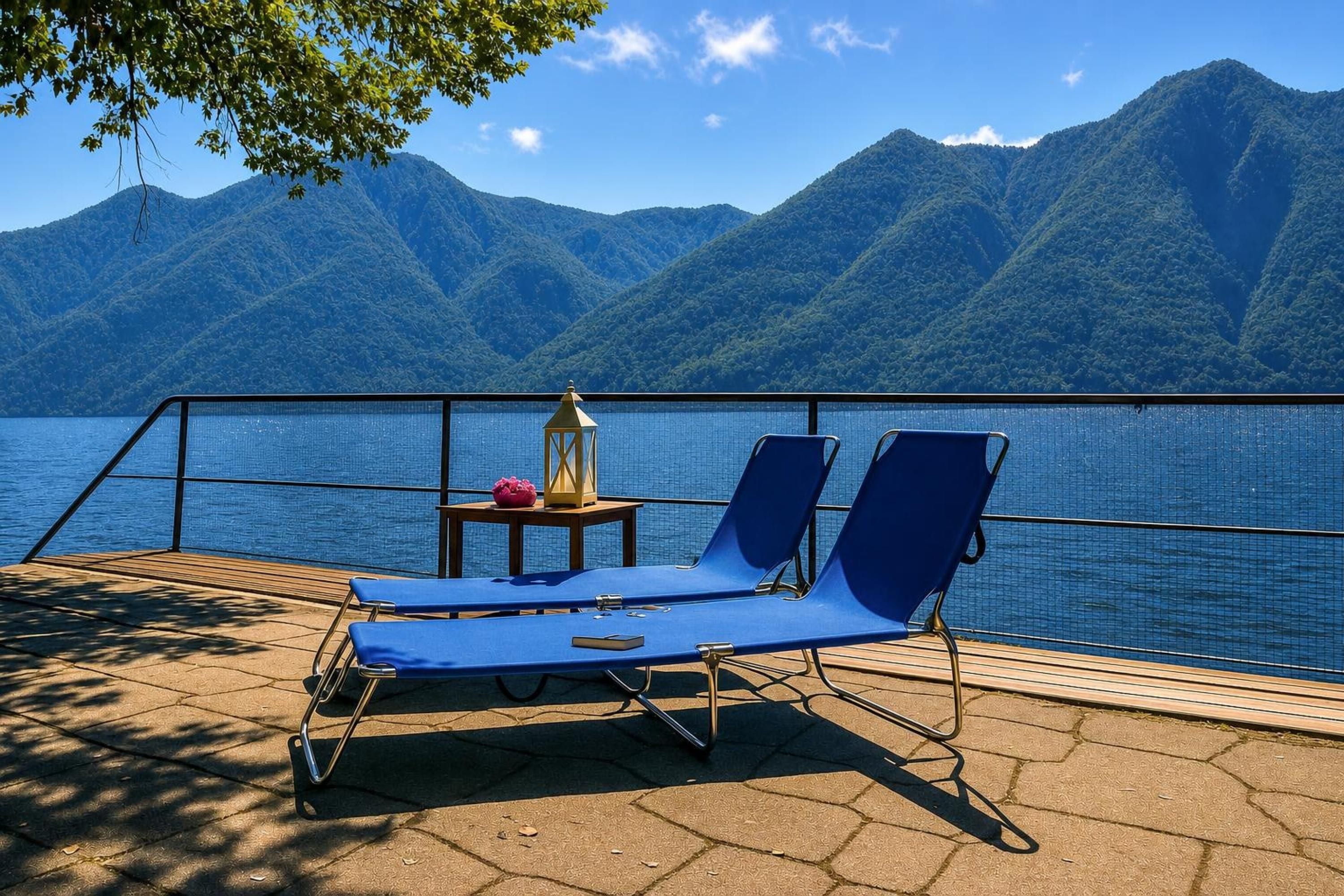 Um terraço com duas cadeiras de descanso azuis, uma mesinha com um candeeiro e vista para um lago com montanhas ao fundo.