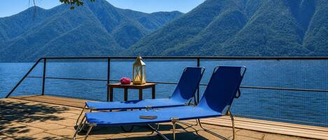 Um terraço com duas cadeiras de descanso azuis, uma mesinha com um candeeiro e vista para um lago com montanhas ao fundo.