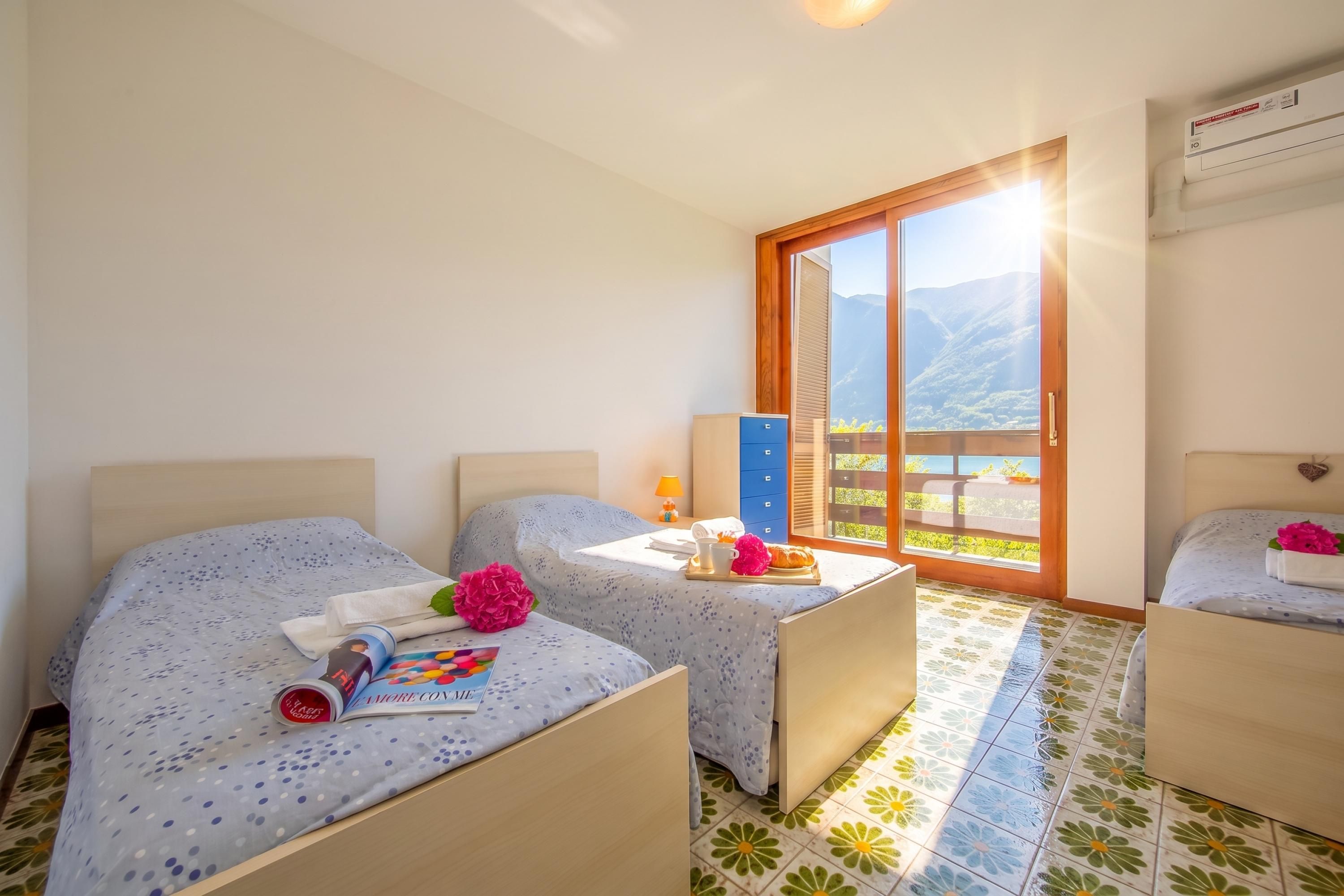 A hotel room with two beds, a small table with a tray of breakfast, a window with a mountain view, and an air conditioning unit.