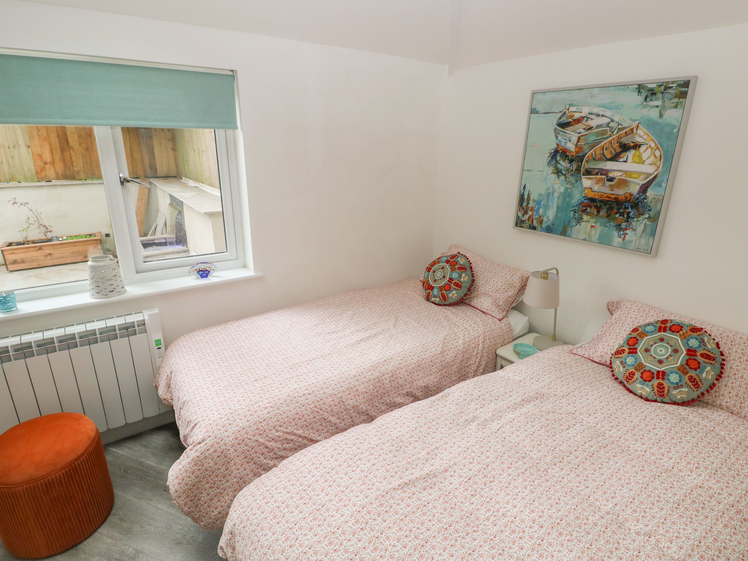 A bedroom with two beds, a window with a view of boats, a radiator, and a small orange stool.