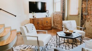 A modern living room with a staircase, a flat-screen TV, wooden cabinets, and a brick wall. - Stunning townhouse on quiet street in Springfield! (Jacksonville)