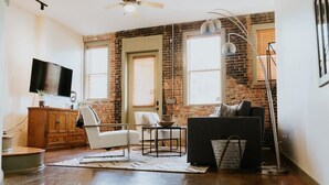 A living room with a brick wall, a flat-screen TV, a white sofa, a black sofa, a small table, and a floor lamp. - Stunning townhouse on quiet street in Springfield! (Jacksonville)