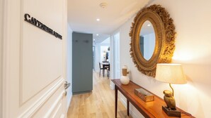 A hallway with a sign reading "CAVENDISH SQUARE," a round mirror with a frayed edge, a wooden console table with a lamp, and a vase.