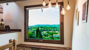 Ein Zimmer mit großem Fenster, das auf eine grüne Landschaft blickt, einer Steinsitzbank und einem Holzboden mit einer Vase und einem Buch.