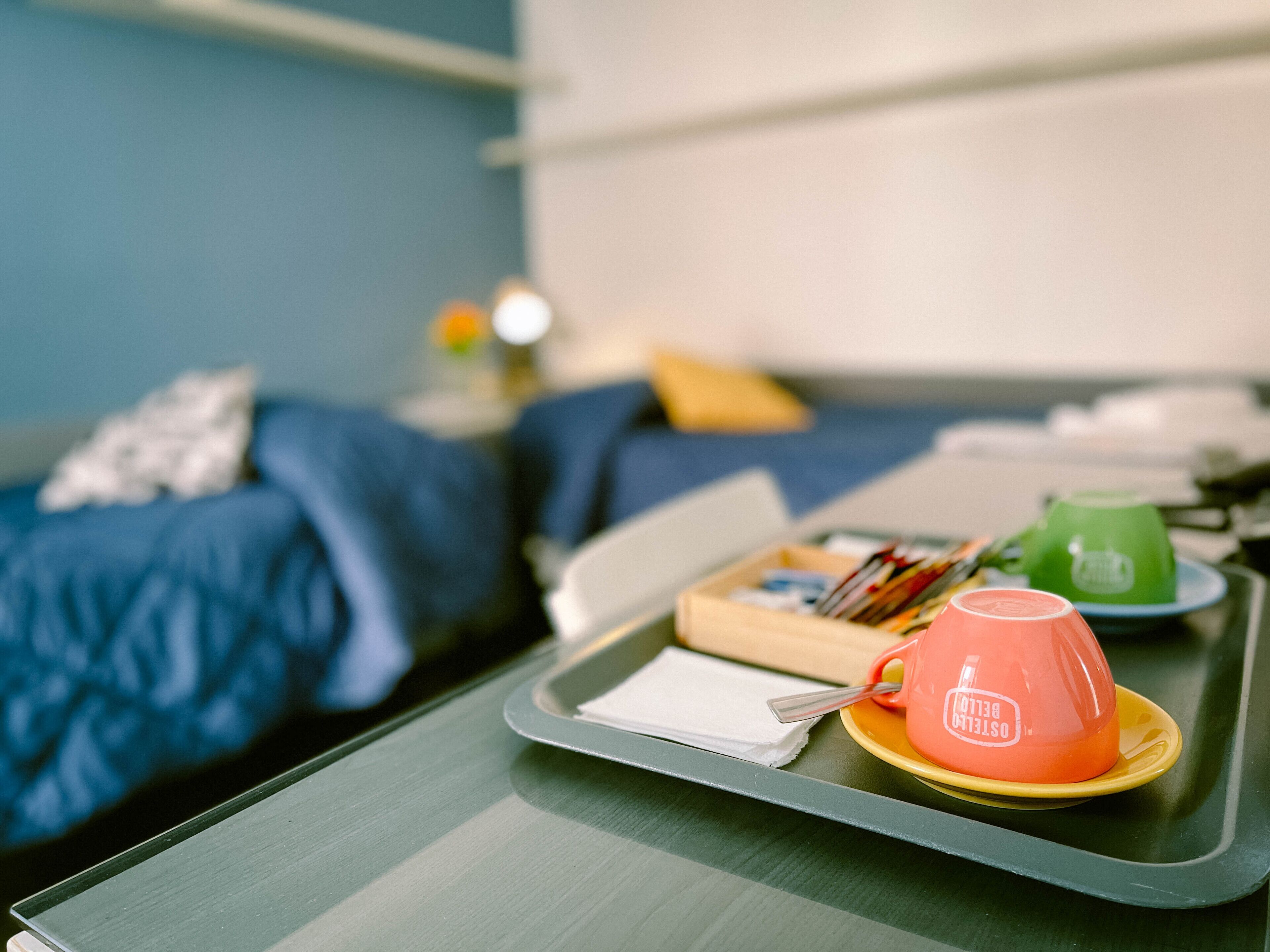 A tray with a pink mug labeled "Osteria," a green cup, and a book on a table in a room with a bed and a blue wall.
