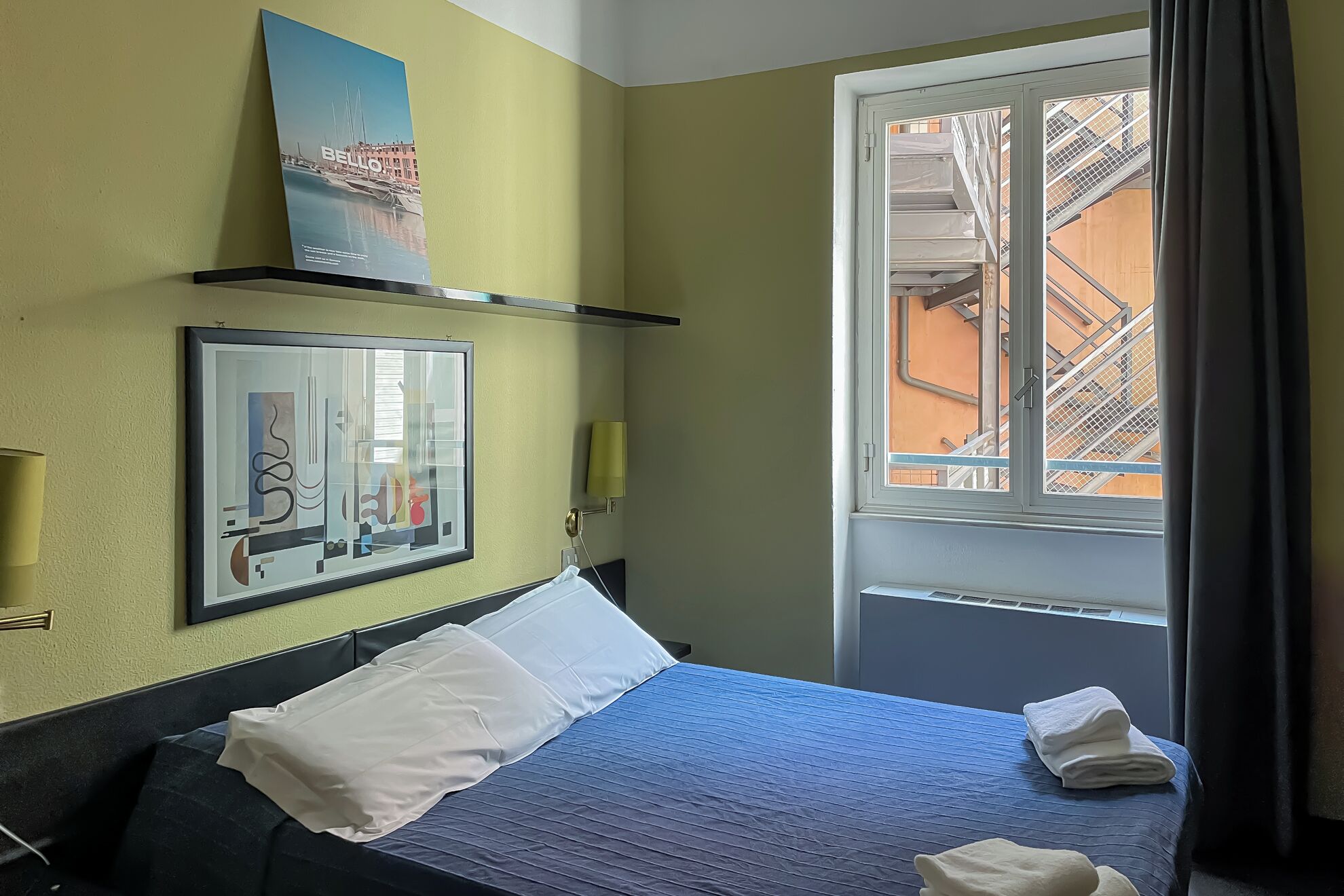 A hotel room with a bed, a window with a view of buildings, a framed picture on the wall, and a shelf with a book.