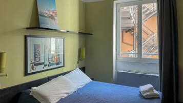 A hotel room with a bed, a window with a view of buildings, a framed picture on the wall, and a shelf with a book.