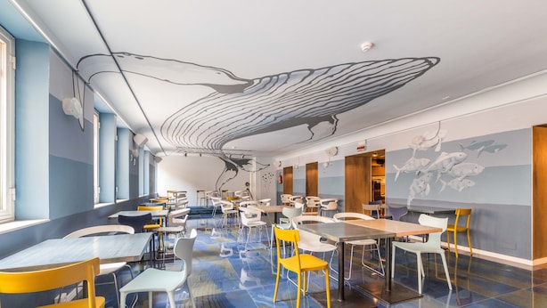 A modern dining area with tables and chairs, a mural on the wall, and a ceiling with a large bird design. - Ostello Bello Genova (Genoa)