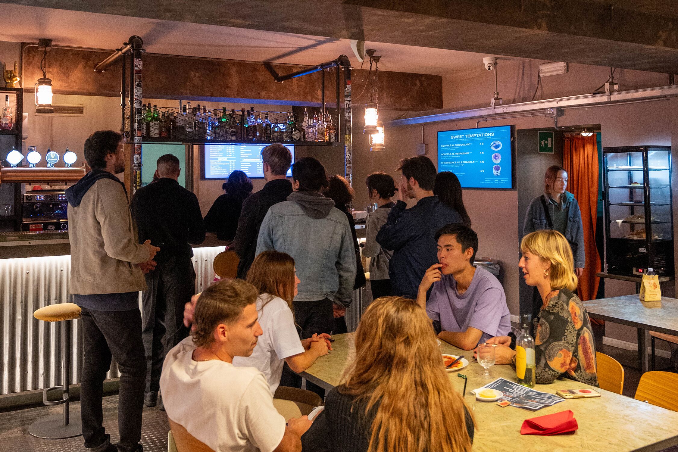 A bar area with a menu board, bar stools, and a group of people seated at tables.