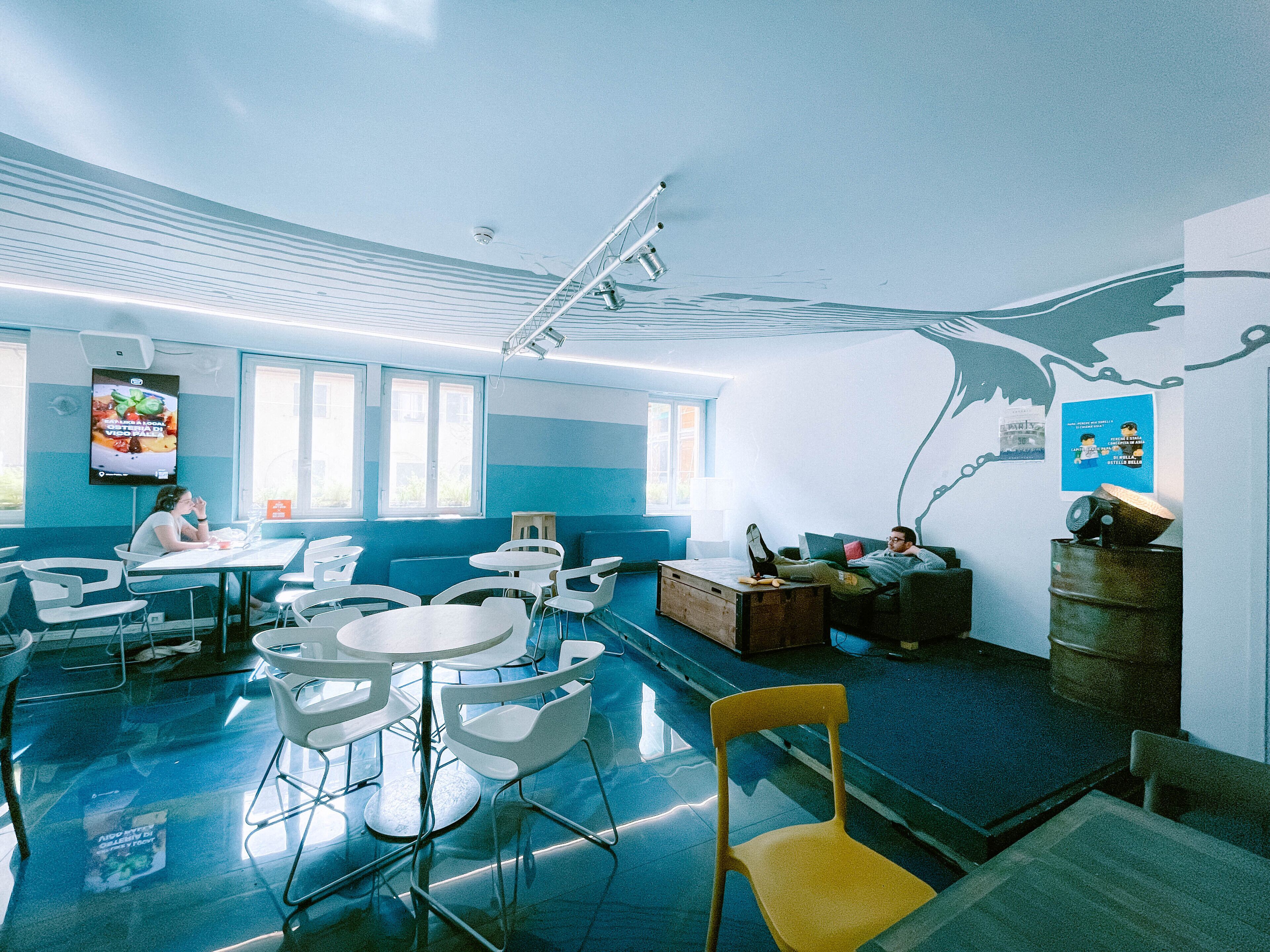 A modern, well-lit room with a curved ceiling, white and blue walls, and a large mural. There are several white chairs and tables, and a person is sitting at a table.