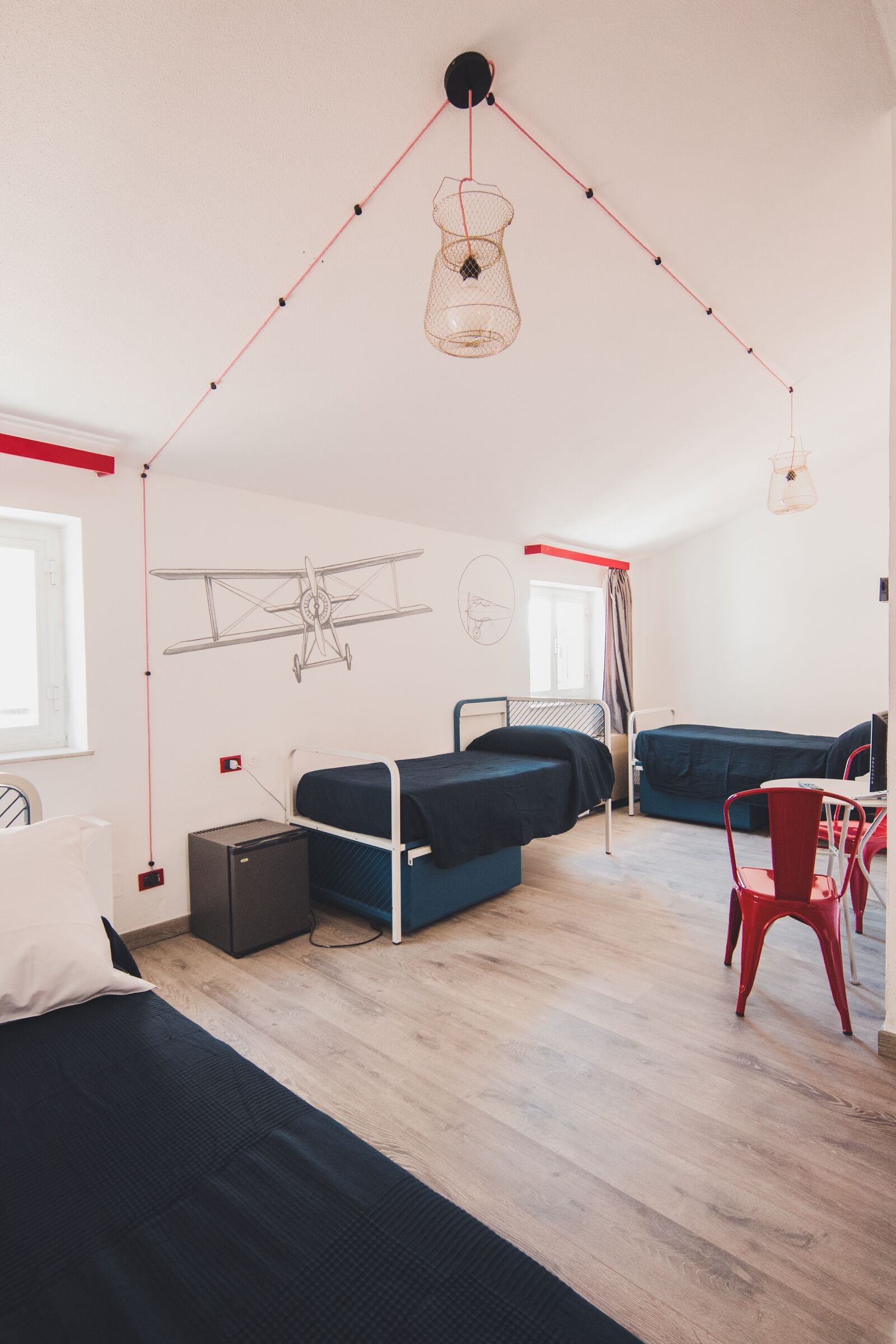 Habitación de residencia estudiantil con literas, un escritorio y una silla. Se ve una ventana, un radiador y una lámpara fijada a la pared.