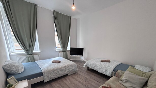 A modern, well-lit room with two beds, a sofa, a TV, and a small table.