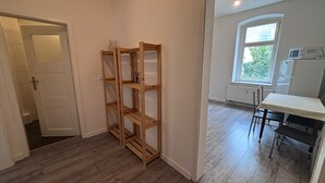 A room with a wooden bookshelf, a white door, a window with a view of greenery, and a kitchen area with a refrigerator and microwave.