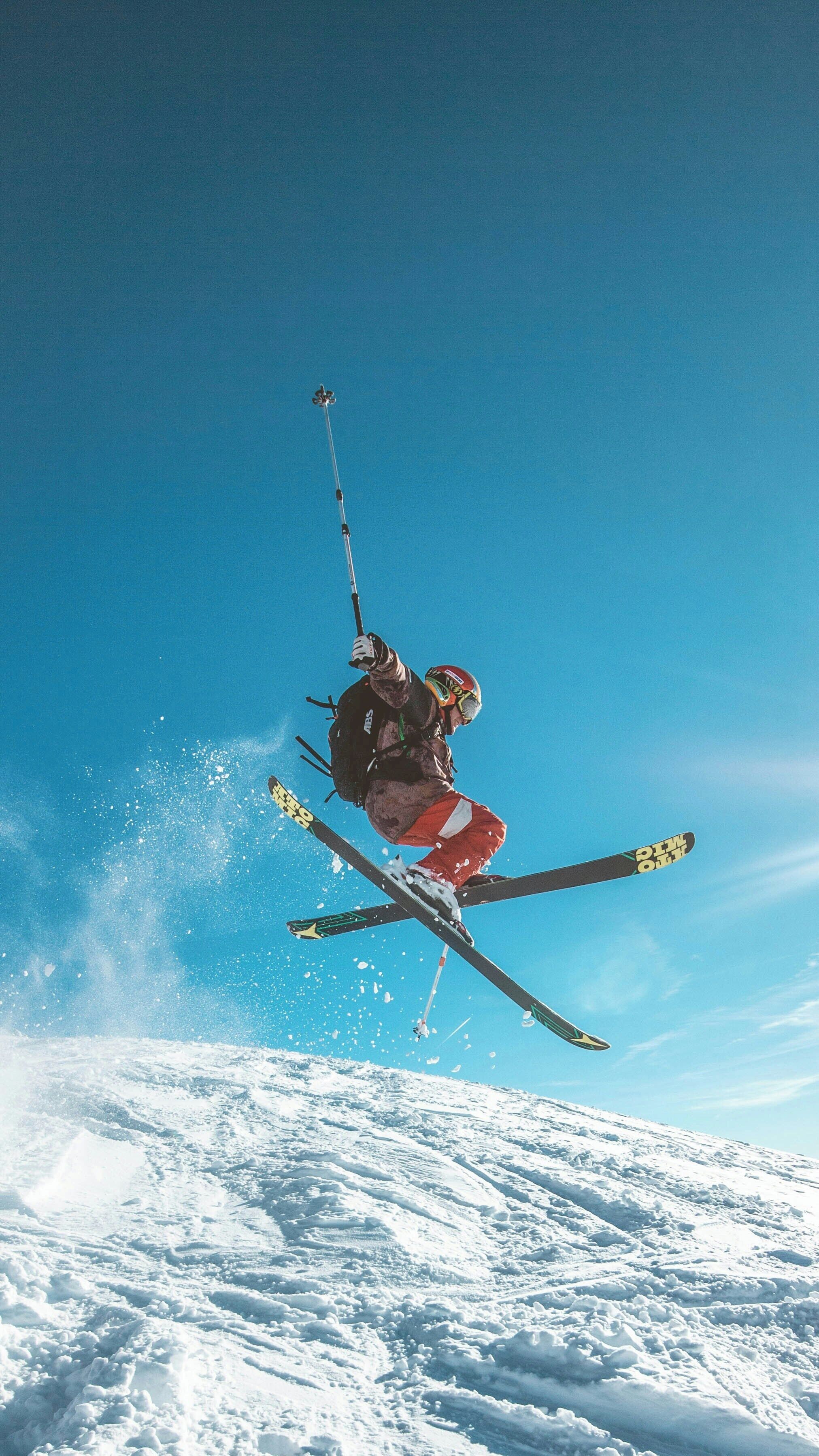 Un skieur en plein saut, en l’air, avec ses skis et ses bâtons, sur fond de ciel bleu dégagé.