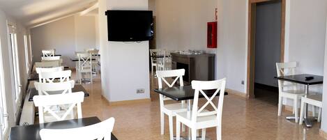 Un espace repas comprenant plusieurs tables et chaises, un téléviseur à écran plat fixé au mur, et un coin cuisine équipé d’un réfrigérateur et d’une cuisinière.
