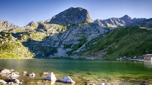 Paisaje montañoso con un lago cristalino, orillas rocosas y un pequeño edificio cerca del agua.