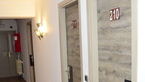 Un pasillo con piso de madera, puertas con los números 211 y 210, y un extintor rojo en la pared.