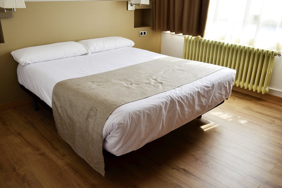 Een netjes opgemaakt bed in een hotelkamer met een houten vloer, een radiator en een raam met gordijnen.