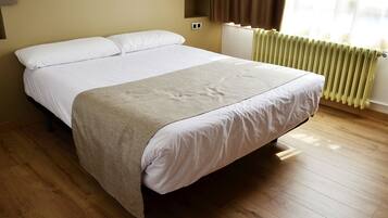 A neatly made bed in a hotel room with a wooden floor, a radiator, and a window with curtains.