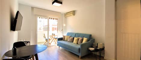 A living room with a blue sofa, a round dining table, and a TV mounted on the wall.