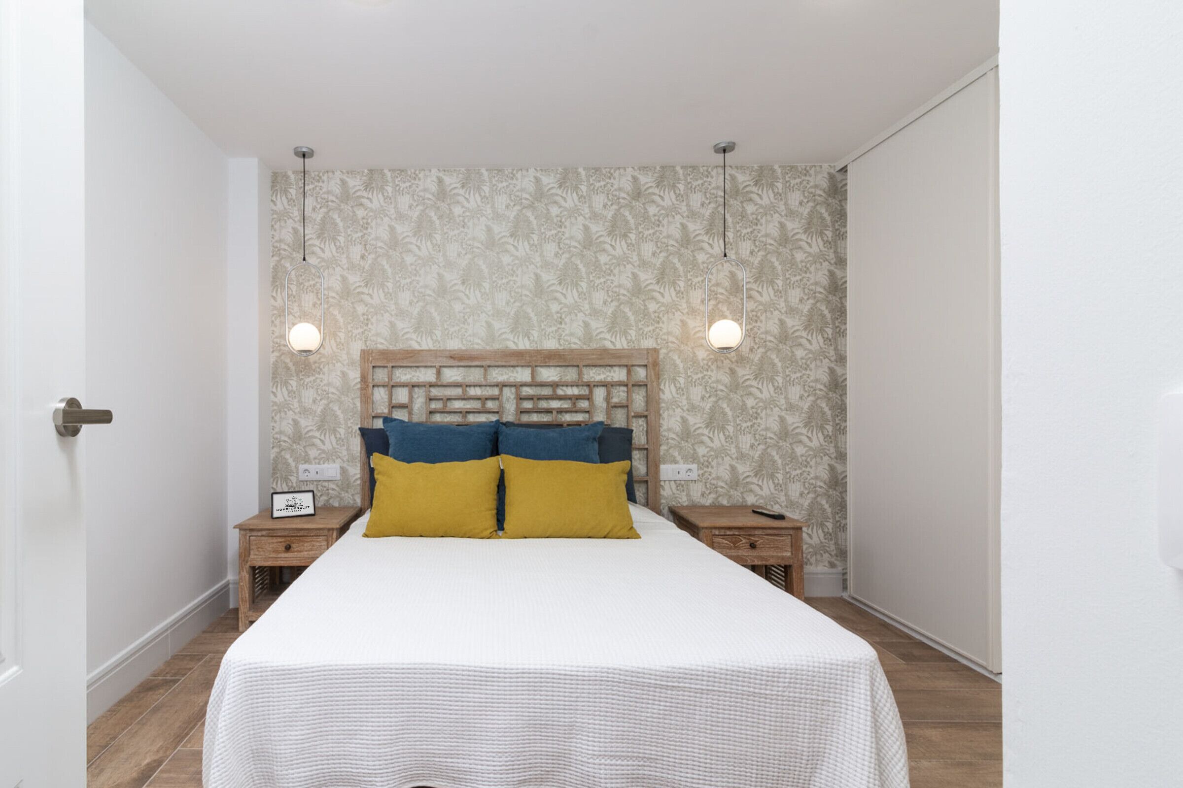 Un dormitorio con cabecera de madera, dos mesitas de noche, una cama con colcha blanca y azul, y dos lámparas colgantes.